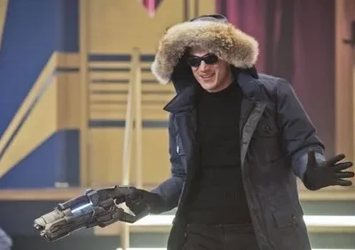 Um homem sorri, vestindo parka com capuz de pele e óculos escuros, segurando uma arma de tecnologia avançada. Fundo de palco colorido.