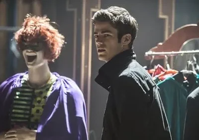 Jovem de jaqueta escura olha surpreso ao lado de manequim com peruca ruiva, máscara, capa roxa, num ambiente escuro com roupas.