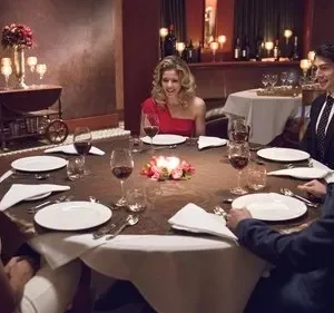 Seis amigos elegantes jantam em restaurante luxuoso à luz de velas. Todos sorriem, conversando. Ambiente aconchegante e festivo.