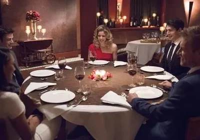 Seis amigos elegantes jantam em restaurante luxuoso à luz de velas. Todos sorriem, conversando. Ambiente aconchegante e festivo.