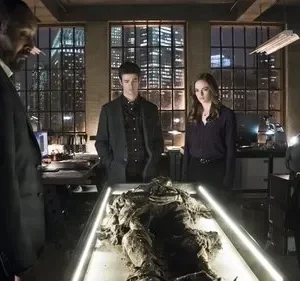 Quatro pessoas sérias em um laboratório urbano à noite, observam um corpo mumificado em uma mesa iluminada. Clima misterioso.