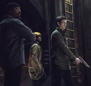 Três homens em um túnel escuro. Um barbudo, um com capacete, e um com jaqueta verde segurando uma arma. Clima sombrio.