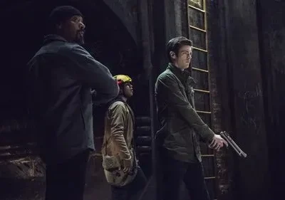 Três homens em um túnel escuro. Um barbudo, um com capacete, e um com jaqueta verde segurando uma arma. Clima sombrio.