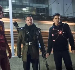 Três heróis de pé: um em traje vermelho com um raio, um arqueiro de casaco escuro, e um de preto com um símbolo luminoso. Cidade à noite.