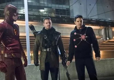 Três heróis de pé: um em traje vermelho com um raio, um arqueiro de casaco escuro, e um de preto com um símbolo luminoso. Cidade à noite.