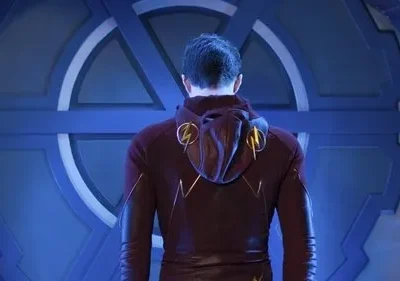 Homem de costas com traje vermelho e raios dourados, em frente a uma imponente porta circular azul de design futurista.