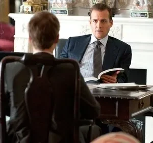 Dois homens em ternos conversam num escritório elegante. Um, sério, segura um livro; o outro está de costas. Clima profissional.