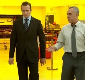 Dois homens em um laboratório industrial amarelo vibrante. Um de terno escuro caminha, enquanto o outro, de camisa e gravata, gesticula.