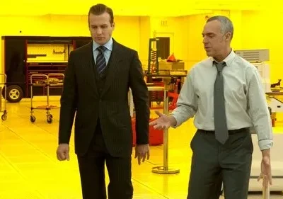 Dois homens em um laboratório industrial amarelo vibrante. Um de terno escuro caminha, enquanto o outro, de camisa e gravata, gesticula.