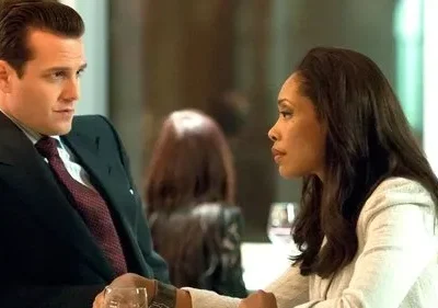 Homem de terno e gravata, e mulher de cabelos longos e blazer claro, trocam olhares sérios num ambiente moderno.