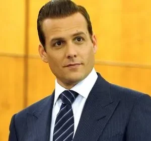 Homem de terno risca de giz, óculos e cabelo penteado, olha com expressão confiante. Fundo profissional amadeirado.