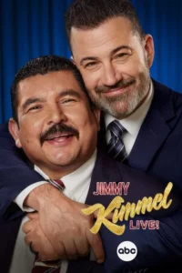 Dois homens sorridentes. Um,de barba,abraça por trás o outro,de bigode. Ambos vestem terno e gravata,em fundo azul.