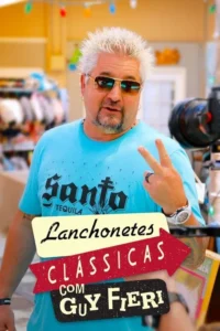 Homem de cabelo branco espetado, óculos escuros e camiseta azul, faz sinal de paz. Fundo colorido e câmera. Clima vibrante.