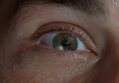 Close-up de um olho com íris verde-acastanhada. Um planeta azul é refletido na pupila, criando um clima intrigante.