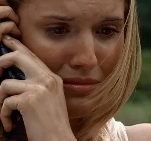 Mulher loira suja com expressão angustiada, falando ao telefone, com floresta desfocada ao fundo.