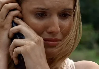 Mulher loira suja com expressão angustiada, falando ao telefone, com floresta desfocada ao fundo.