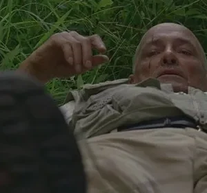 Homem careca com cortes, deitado na grama, olhando para cima. Veste roupa clara; uma bota em primeiro plano. Clima de exaustão.