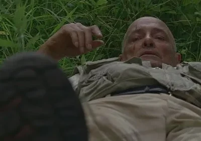 Homem careca com cortes, deitado na grama, olhando para cima. Veste roupa clara; uma bota em primeiro plano. Clima de exaustão.