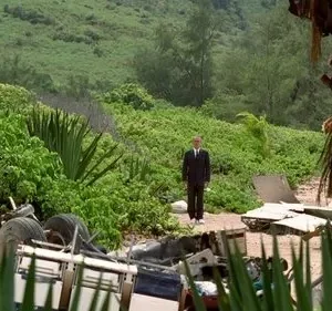 Homem de terno parado em meio a uma paisagem tropical exuberante, com destroços no primeiro plano e folhagem densa. Ambiente estranho e isolado.