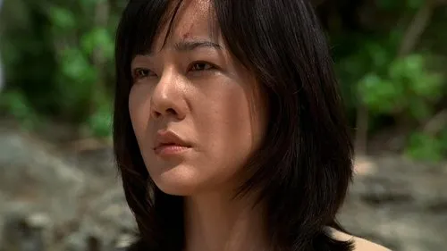 Uma mulher de cabelo escuro,com um corte na testa,olha séria. Fundo desfocado de folhagem verde e rochas. Clima pensive.