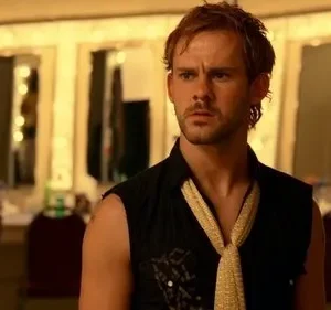 Homem ruivo com barba e camisa preta com gravata dourada em camarim iluminado, olhando pensativo.