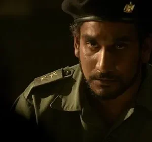 Um homem barbudo de uniforme militar verde-oliva e boina preta, com olhar intenso, emerge da escuridão. Estrelas nas ombreiras.