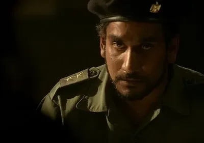 Um homem barbudo de uniforme militar verde-oliva e boina preta, com olhar intenso, emerge da escuridão. Estrelas nas ombreiras.