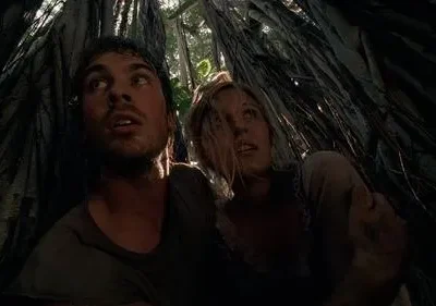 Homem e mulher olham apreensivos para cima, cercados por raízes escuras em uma selva densa. Luz do alto filtra o clima tenso.