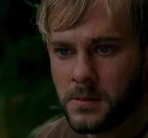 Close-up de um homem barbudo loiro de olhos azuis, com expressão séria, em meio a folhagem verde borrada.