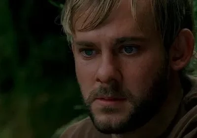 Close-up de um homem barbudo loiro de olhos azuis, com expressão séria, em meio a folhagem verde borrada.