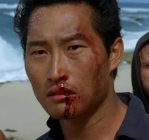 Close-up de homem asiático com rosto ensanguentado e sujo, olhar intenso. Dois homens desfocados na praia ao fundo. Clima tenso.