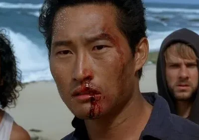 Close-up de homem asiático com rosto ensanguentado e sujo, olhar intenso. Dois homens desfocados na praia ao fundo. Clima tenso.