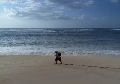 Uma pessoa caminha sozinha na areia clara de uma praia vasta. O oceano azul tem ondas quebrando, e há nuvens no céu. Pegadas na areia.