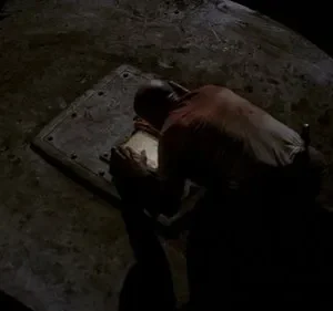 Homem careca ajoelhado em chão escuro, focado num painel retangular luminoso. Cenário sombrio e misterioso.