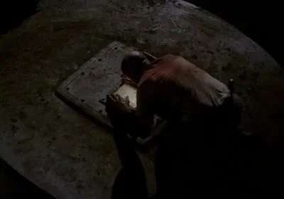 Homem careca ajoelhado em chão escuro, focado num painel retangular luminoso. Cenário sombrio e misterioso.