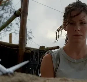 Mulher séria, cabelo úmido, olha à frente. Cenário rústico com estrutura de bambu. Um modelo de avião no chão. Clima de incerteza.