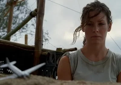 Mulher séria, cabelo úmido, olha à frente. Cenário rústico com estrutura de bambu. Um modelo de avião no chão. Clima de incerteza.