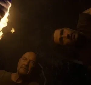 Pôster escuro. Um homem careca e outro jovem olham para cima, iluminados por uma tocha flamejante. Atmosfera tensa e misteriosa.