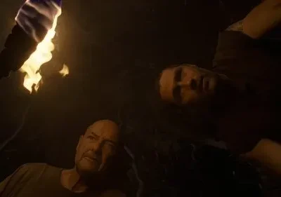 Pôster escuro. Um homem careca e outro jovem olham para cima, iluminados por uma tocha flamejante. Atmosfera tensa e misteriosa.