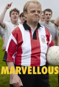 Um homem loiro de meia-idade com camisa de futebol vermelha e branca, segurando uma bola. Ele tem um sorriso confiante, com jovens num campo verde.