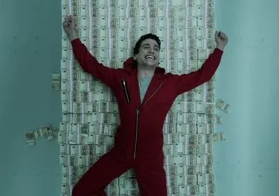 Jovem de macacão vermelho, sorrindo vitorioso, deitado sobre parede de notas de euro dentro de um cofre azul.