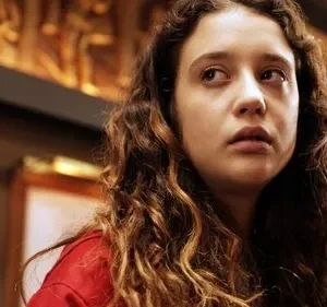 Jovem com cabelo cacheado, expressão séria, olha para cima. Veste vermelho, fundo interno com detalhes dourados.