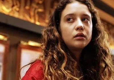 Jovem com cabelo cacheado, expressão séria, olha para cima. Veste vermelho, fundo interno com detalhes dourados.