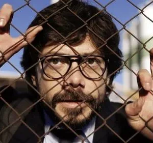 Homem de óculos e barba segura grade enferrujada, olhando fixamente. Céu azul e prédio industrial ao fundo, criando clima tenso.