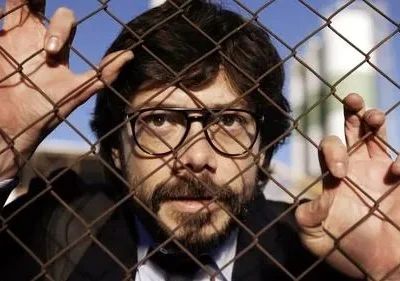 Homem de óculos e barba segura grade enferrujada, olhando fixamente. Céu azul e prédio industrial ao fundo, criando clima tenso.