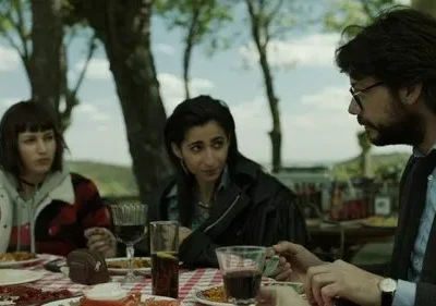 Três pessoas à mesa de piquenique: um homem de óculos e barba e duas mulheres, uma de xadrez. Conversam seriamente ao ar livre.