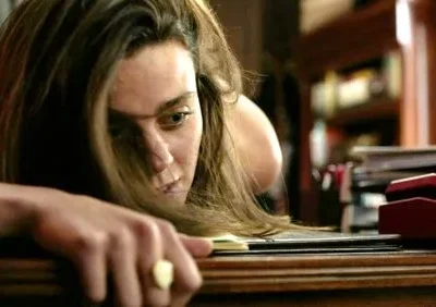 Mulher com cabelos soltos e rosto cansado, apoiada numa mesa de madeira com anel no dedo. Fundo desfocado com livros e papéis. Clima melancólico.