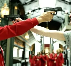 Dois homens se confrontam, ambos apontando armas para a cabeça um do outro. Um sorri, o outro grita. Pessoas de macacão vermelho ao fundo.