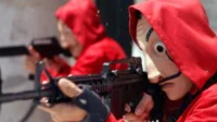 Uma figura em macacão vermelho e máscara surreal com bigodes e olhos arregalados mira um rifle. Outra figura similar desfocada ao fundo. Tensão.