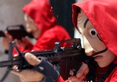 Uma figura em macacão vermelho e máscara surreal com bigodes e olhos arregalados mira um rifle. Outra figura similar desfocada ao fundo. Tensão.